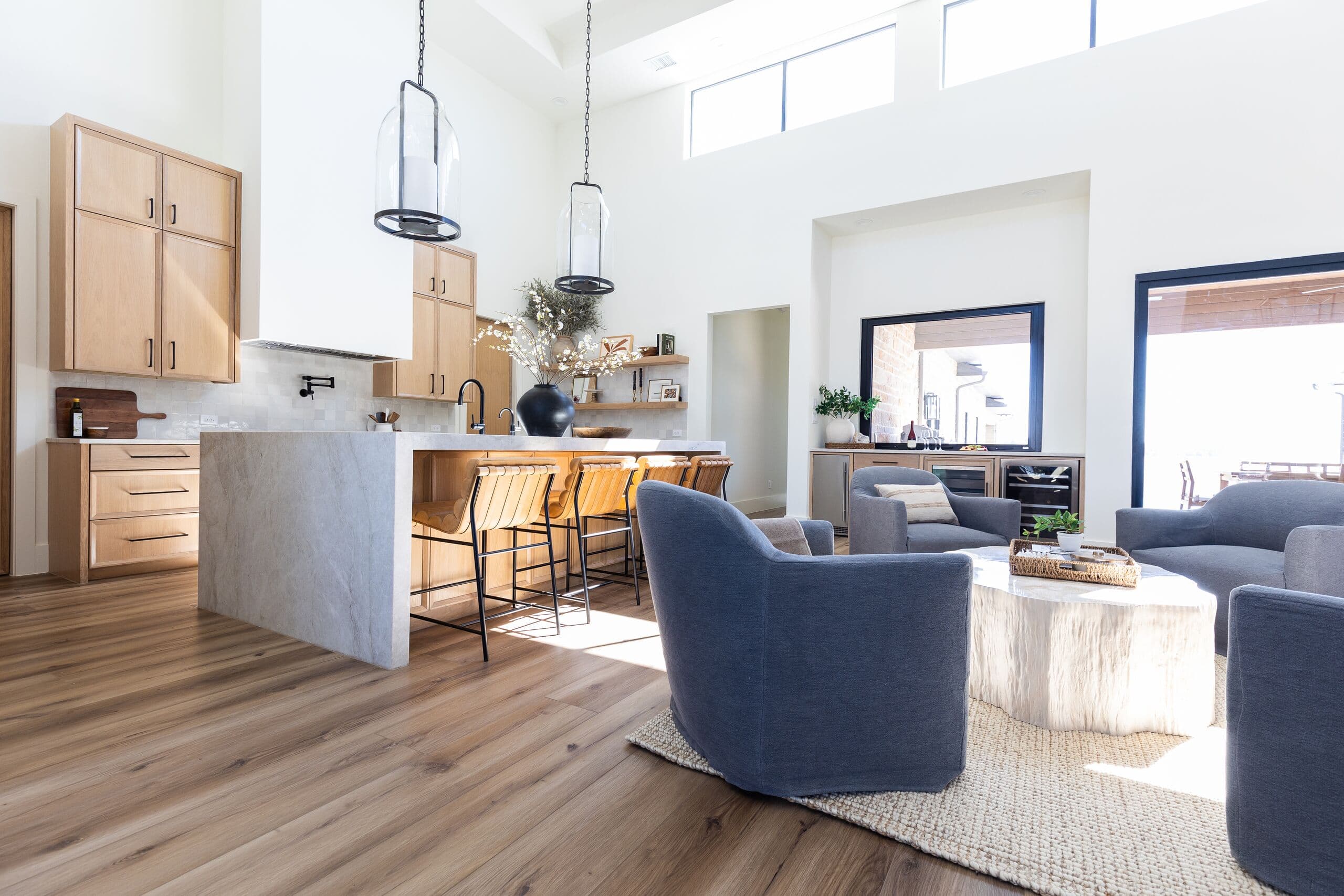 Open kitchen and living area with a stone island, leather stools, blue slipcovered swivel chairs, a pale carved coffee table and tall windows.